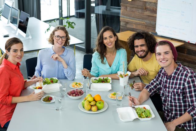 Diverse group of business professionals enjoying a healthy lunch together in a modern office environment. Ideal for use in articles or advertisements about workplace culture, team building, corporate wellness programs, or promoting healthy eating habits in the office.