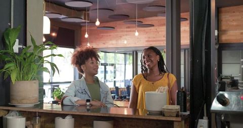 Two women chatting at cozy cafe counter with potted plant and natural light
