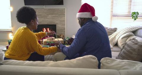 Couple wrapping christmas gifts by fireplace wearing santa hat and mustard sweater