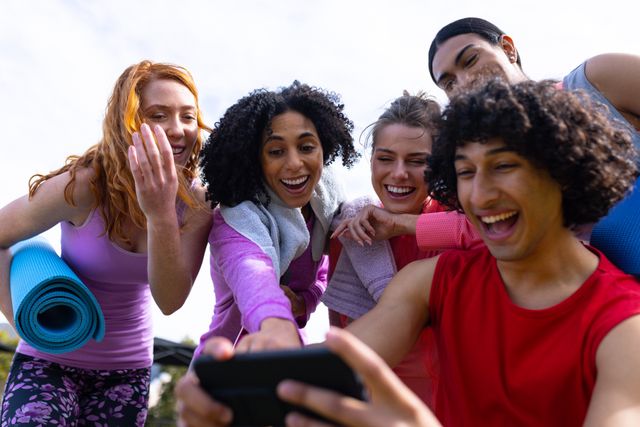 Diverse group of friends laughing and looking at a smartphone after a yoga session outdoors. Perfect for promoting healthy lifestyle, fitness, friendship, and outdoor activities. Ideal for use in advertisements, social media campaigns, and wellness blogs.