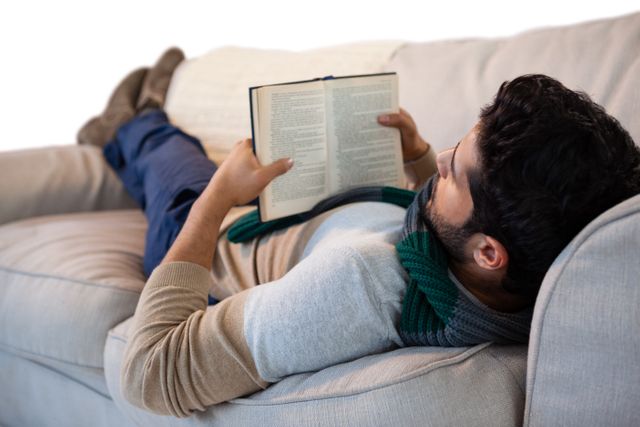 Man lying on a sofa while reading a book, wearing casual clothes and a scarf. Ideal for use in articles or advertisements related to relaxation, leisure activities, home lifestyle, or reading habits. Perfect for illustrating concepts of comfort and personal time.