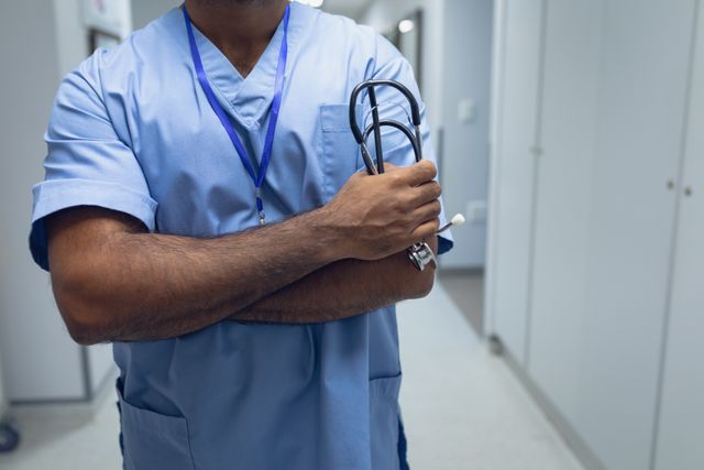 Mid section of biracial male doctor standing with arms crossed in the corridor at hospital
