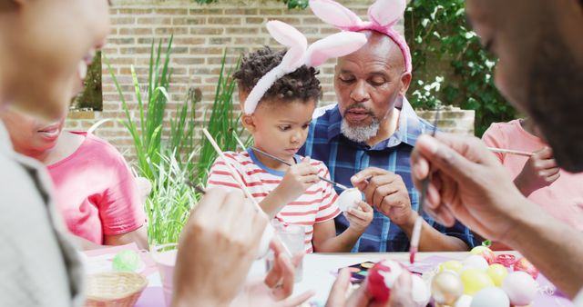 Multi-generational family sitting outside, painting Easter eggs, enjoying quality time. Grandpa helping child decorate, everyone involved in festive activity. This can be used for holiday promotions, family-themed advertisements, or articles about bonding activities.