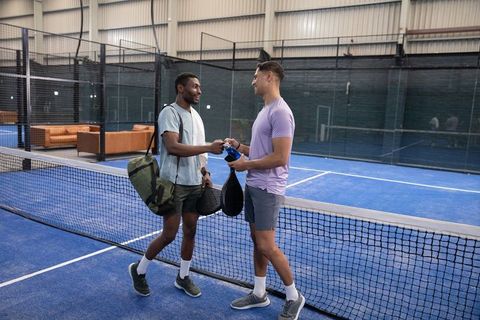 Two men shaking hands and passing water bottle on blue padel court