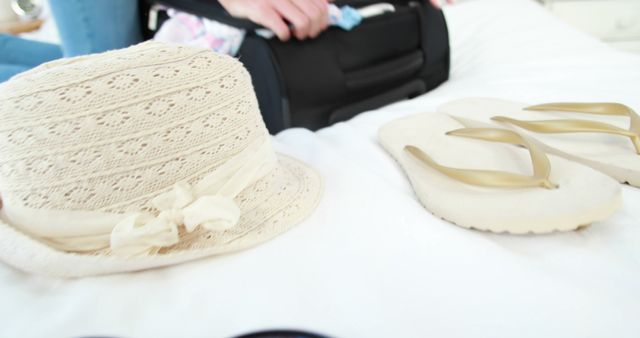 A straw hat and a pair of flip-flops are in focus, suggesting a vacation or beach setting, with copy space. Out of focus in the background, a person appears to be packing or unpacking a suitcase, emphasizing travel preparation or the end of a trip.