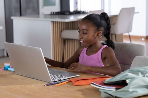 African american girl studying online at kitchen table with laptop and notebooks
