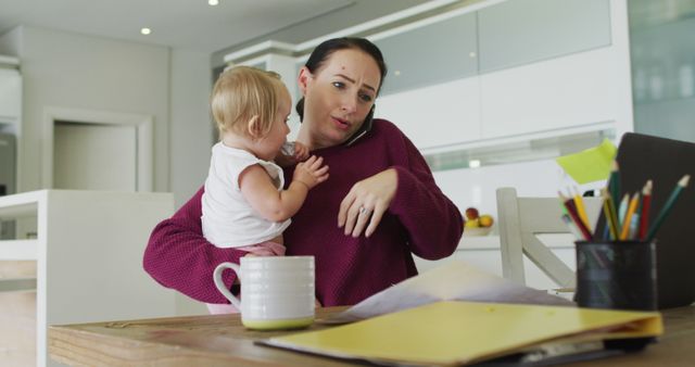 Caucasian mother holding her baby and talking on smartphone while working from home. motherhood, love and childcare concept