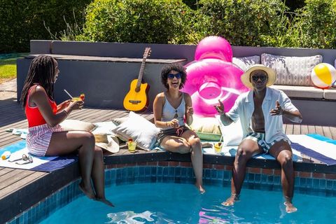 African friends laughing poolside with summer drinks, guitar, sunglasses and pink float
