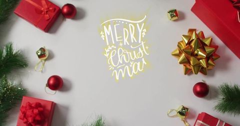 Hand-lettered gold merry christmas flatlay with red gifts, baubles, gold bow and evergreen sprigs