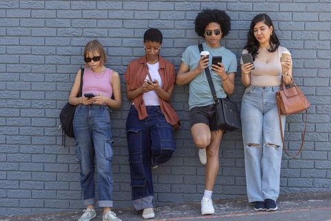 Diverse friends leaning on blue brick wall checking smartphones and holding coffee cups