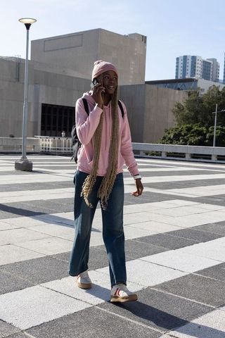 African american woman walking and talking on smartphone in urban plaza wearing pink hoodie