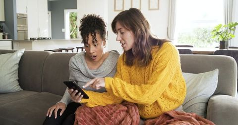 Diverse mid-adult women sitting on gray couch pointing at smartphone, sharing surprised cozy moment