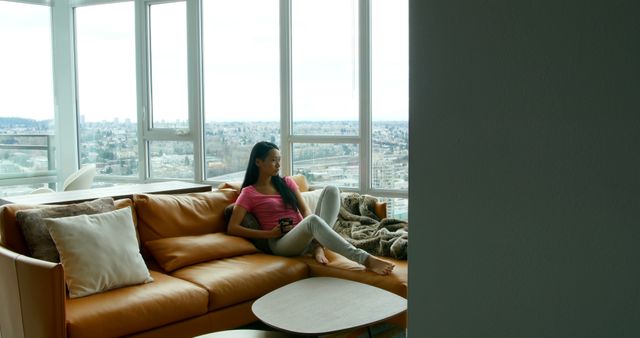 Young woman relaxing on sofa in modern high-rise apartment with city view - Download Free Stock Images Pikwizard.com