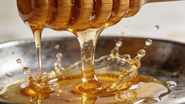 Drizzling honey from wooden dipper forming golden splashes and glossy droplets, macro