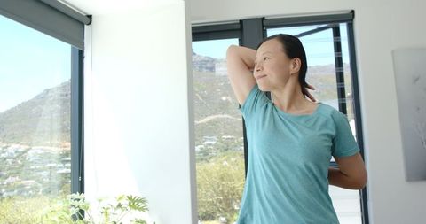 Asian woman stretching by sunlit window in modern apartment for wellness and relaxation
