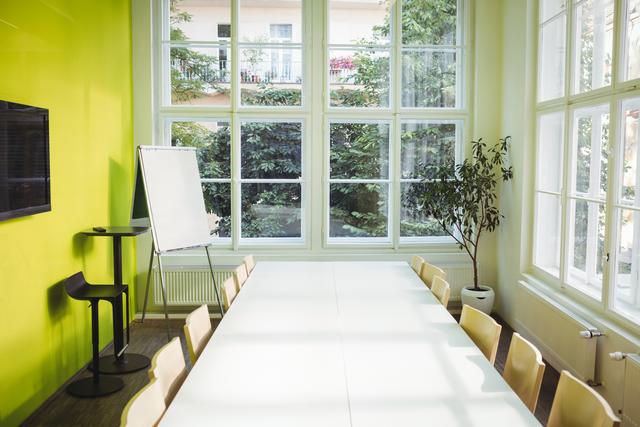 Bright modern office meeting room featuring large windows, conference table, chairs, whiteboard, and indoor plant. Ideal for business presentations, corporate meetings, and professional workspace settings.