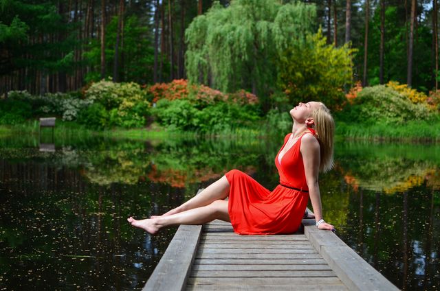 Woman in orange dress sitting on dock by tranquil lake surrounded by lush green forest. She is barefaced and appears relaxed enjoying nature. This image captures peaceful outdoor recreation and can be used for wellness, relaxation, travel, nature retreats, or summer getaway promotions.