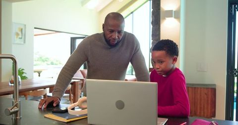 African american father and son leaning on laptop at kitchen island, studying together
