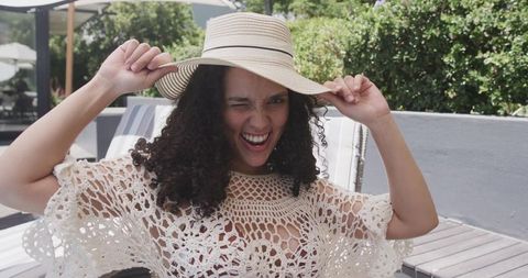 African american woman winking while holding straw hat on sunny poolside lounge chair