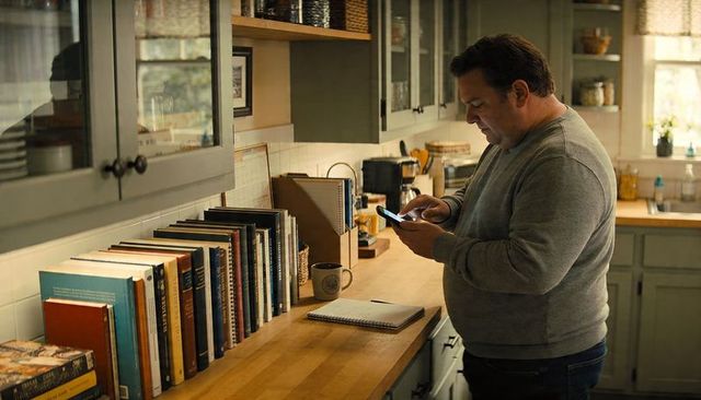 Middle-aged man checking smartphone in cozy home kitchen lined with books and notebook
