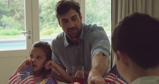 Father spending quality time with his two children at home, celebrating Independence Day. He is handing something to his kids, who are holding American flags. Ideal for content related to family gatherings, holiday celebrations, patriotism, and Independence Day activities.