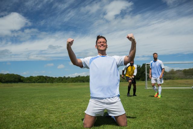 Excited football player in celebrating scoring goal kneeling on grass pitch on a sunny day