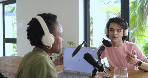 Diverse duo recording podcast in bright home studio with laptop and mics, sunlit greenery