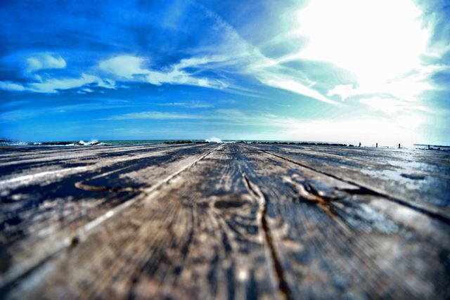 Wide-angle view of a rustic wooden pier leading towards the ocean under a bright, sunny sky. Ideal for travel websites, inspirational quotes, relaxation themes, and seaside adventure promotions.