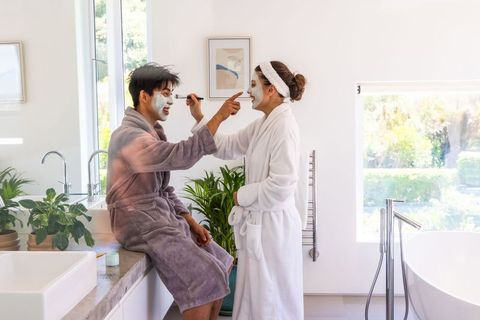 Couple sharing at-home spa moment, applying facial masks and smiling in sunlit bathroom