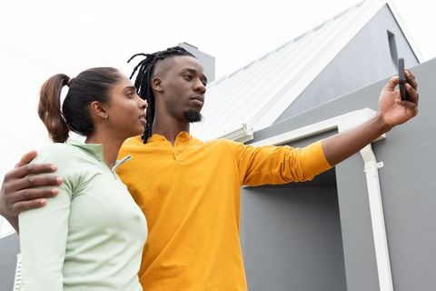 Young couple taking selfie outside modern grey house embracing during overcast day