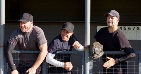 Three baseball teammates cheering in dugout, leaning on fence with glove and scoreboard