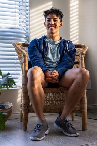 Sunlit asian young man sitting in rattan armchair smiling in minimalist home corner