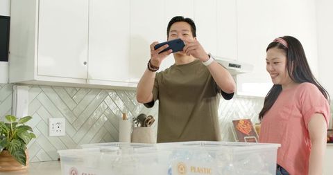 Asian couple recording on phone sorting kitchen recycling bins labeled plastic at home