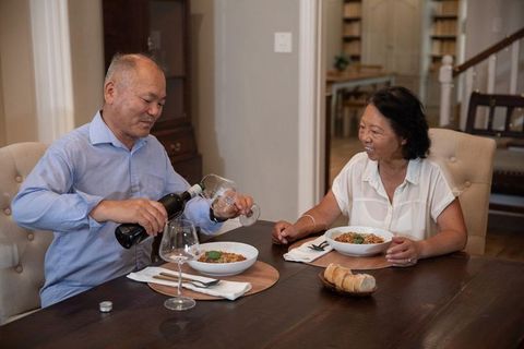 Asian couple sharing cozy homemade dinner at dining table with wine pouring and smiling