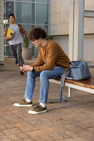 Diverse students sitting on campus bench using smartphone while classmate walking with folder