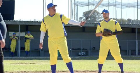 Mature Batter Swinging Bat in Yellow Baseball Uniform with Teammate in Dugout