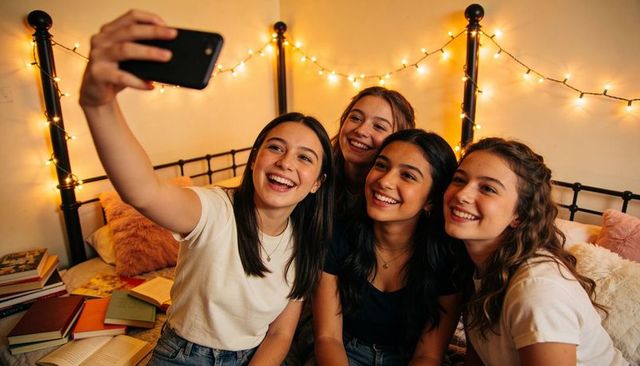 Teenage friends taking selfie on bed, laughing together in cozy bedroom with string lights
