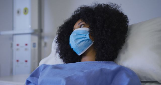Young woman with curly hair wearing a face mask, lying on hospital bed, looking off the frame. Suitable for use in healthcare, medical treatment, hospital care, recovery process themes, patient safety, and protective measures. Useful for articles related to healthcare systems, patient care, and safety protocols.