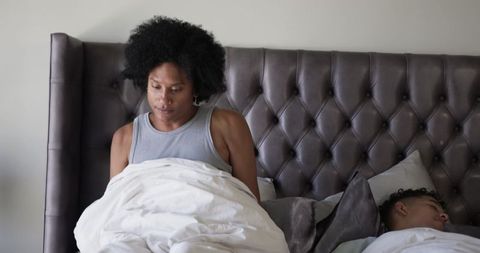 Pensive partner sitting upright in bed gazing away while partner sleeping on tufted headboard