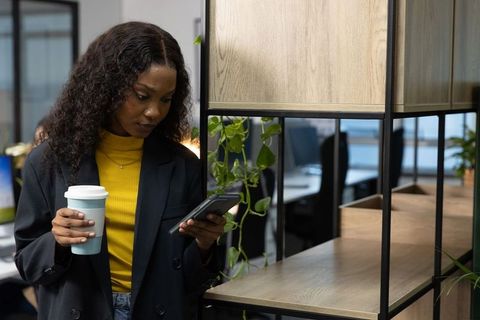 African american professional checking phone while holding coffee in modern open-plan office