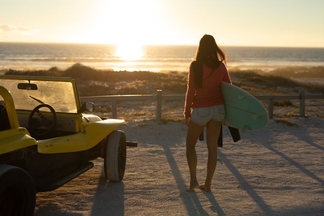 Caucasian woman standing by a yellow beach buggy on a sandy beach at sunset, holding a surfboard and admiring the ocean view. Ideal for themes of summer holidays, road trips, adventure, relaxation, and outdoor lifestyle. Perfect for travel blogs, vacation advertisements, and lifestyle magazines.