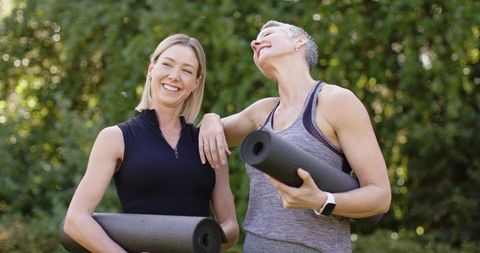 Women laughing while carrying yoga mats in sunny park, activewear wellness lifestyle