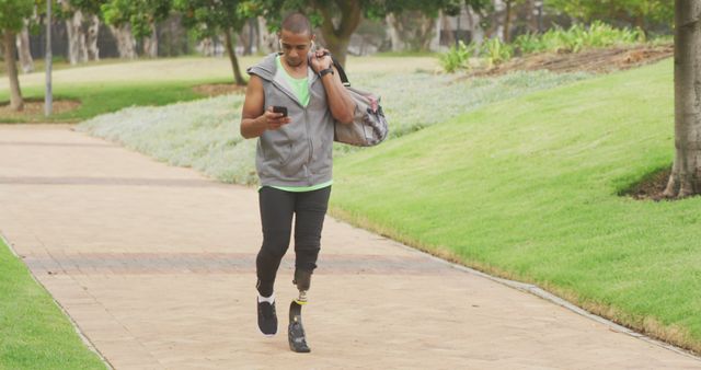 Image shows a young man wearing athletic clothing and carrying a gym bag, walking through a park while looking at his phone. He has a prosthetic leg, which illustrates strength, determination, and active lifestyle. Beautiful green grass and trees in the background, indicating a pleasant, sunny day. Perfect for use in promoting inclusivity in sports, health and wellness campaigns, outdoor activity advertisements, and athletic product promotions.