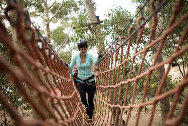 Woman walking on a rope bridge in an adventure park, wearing safety gear and helmet. Ideal for use in content related to outdoor activities, adventure parks, fitness, recreation, and nature exploration.