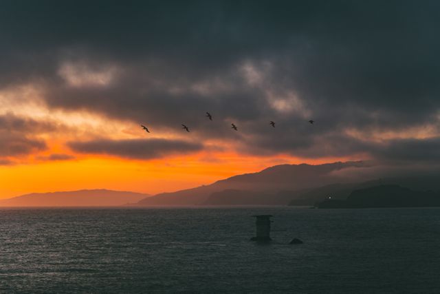 This image shows a serene twilight seascape with vibrant orange hues contrasting dark clouds, and a flock of birds flying above the calm ocean. Perfect for use in travel magazines, nature blogs, and websites highlighting scenic destinations, evening tranquility, or coastal beauty.