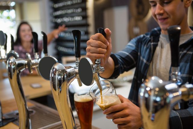 Young bartender pouring beer from tap at modern bar. Ideal for illustrating hospitality industry, bar and pub environments, nightlife, and casual dining settings. Useful for marketing materials, advertisements, and articles related to beverages, service, and social activities.