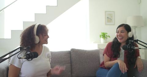 Multicultural women podcast cohosts laughing and chatting on couch with microphones