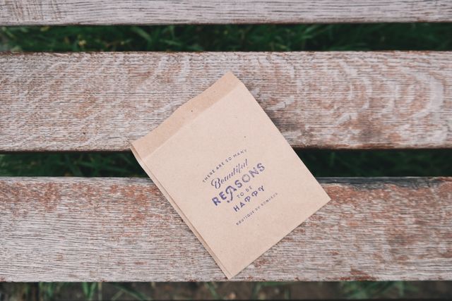 Small beige paper bag with motivational message 'Beautiful Reasons to be Happy' on wooden bench, conveys positivity and encouragement. Ideal for advertisements, blogs about motivation, lifestyle, or nature. Great for use in promotions for eco-friendly products or handcrafts.
