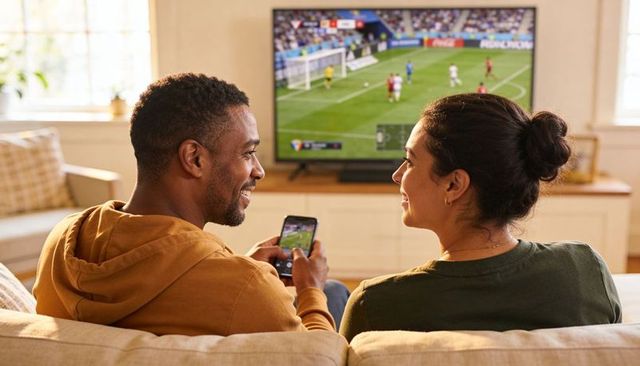 Couple watching live soccer match on tv, chatting and using smartphone on cozy sofa