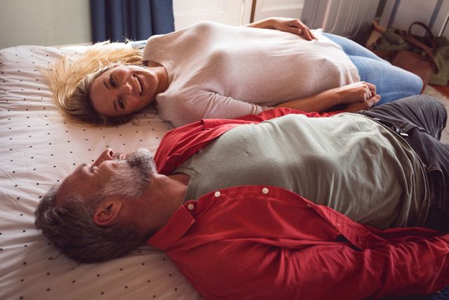 Caucasian couple lying on bed at home, smiling and looking at each other. Perfect for use in advertisements, blogs, or articles about relationships, love, home life, and relaxation.